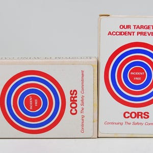May include: Two white rectangular playing card boxes featuring a red and blue bullseye graphic. The text reads OUR TARGET ACCIDENT PREVENTION, INCIDENT FREE, and CORS Continuing The Safety Commitment in red lettering on the front of the boxes.