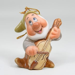 Puede incluir: Una figura de cerámica de un personaje con sombrero beige y camisa gris, tocando un instrumento de cuerda marrón claro con un arco. El personaje tiene una nariz grande, mejillas sonrosadas y un cordón dorado para colgar en el sombrero.
