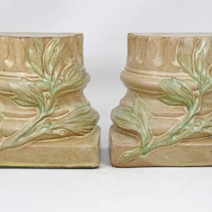 May include: A pair of beige ceramic bookends, each resembling a classical column base. They feature a light green leaf design and sit on a square base. These bookends are a decorative item, suitable for a home.