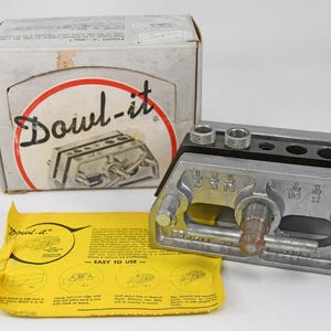 Puede incluir: Una plantilla "Dowl-it" de color plateado con una parte superior negra, acompañada de su caja original y una hoja de instrucciones amarilla. La caja presenta el logotipo "Dowl-it" en un círculo rojo. La herramienta tiene múltiples agujeros y piezas ajustables.