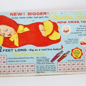 Könnte beinhalten: Vintage Spankin' Sue Puppenwerbung. Das Bild zeigt eine rote Babypuppe mit gelben Details auf hellblauem Hintergrund. Der Text enthält "2 FEET LONG" und Puppenmerkmale. Ein nostalgisches Sammlerstück.