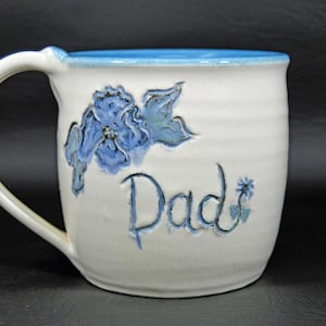 Op de afbeelding: Witte keramische mok met een blauw interieur en handvat. De mok heeft een blauw bloemmotief en het woord "Dad" in blauw schrift. Een klein blauw bloemetje staat naast het woord "Dad".