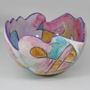 May include: A hand-painted ceramic bowl with a scalloped rim. The bowl features a colorful abstract design with pink, purple, blue, and yellow hues. The rim is outlined in purple.