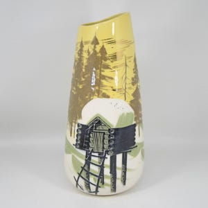 May include: A tall, ceramic vase with a yellow top and a landscape design. The design features a cabin on stilts, a ladder, and a forest scene in shades of brown, green, and black. The vase has a unique, asymmetrical shape.