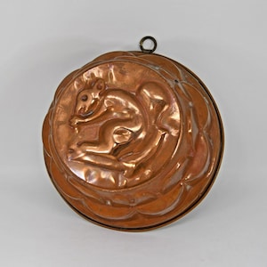 May include: A circular copper mould featuring a squirrel motif. The squirrel is centrally positioned, encircled by an ornate border. A small ring is affixed at the top for suspension. This kitchen utensil is probably used for baking or decorative display.