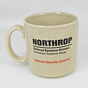May include: A speckled beige ceramic mug with a large handle. The mug features the text "NORTHROP" in black, with additional text in black and red, including "Defense Systems Division" and "Defense Security Systems."