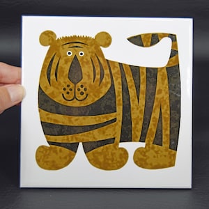 May include: A square ceramic tile featuring a stylized illustration of a tiger against a white background. The tiger is rendered in shades of mustard yellow and dark brown, with a rounded body, a long tail, and a simple face with two eyes.