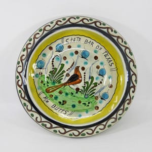 May include: Hand-painted ceramic plate with a bird and floral design. The plate features a yellow center with the text "STATE BAR OF TEXAS" and "M/M BUTTS". The rim has a decorative border with brown and green accents.