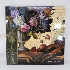 May include: Square decorative box with a floral and musical theme. The box features a still life of flowers, a violin, and the words "Opera House" and "Milbrook". The color palette includes shades of pink, purple, and brown.