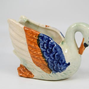 Puede incluir: Jardinera de cerámica con forma de cisne. El cisne tiene un cuerpo blanco, patas y pico naranjas y detalles de alas azules y naranjas. La jardinera tiene un cuello curvado y una pequeña abertura.