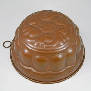 May include: A copper-colored, antique-style Bundt pan with a decorative floral design on the top. The pan has a scalloped edge and a small ring handle. This baking pan is ideal for making cakes and desserts.