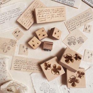 May include: A collection of wooden stamps with various designs, including script, stars, and floral patterns. The stamps are arranged with stamped impressions on aged paper, creating a vintage aesthetic. The image includes handwritten notes and a lace trim.