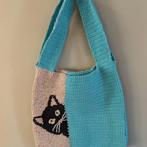 May include: A handmade crochet tote bag with a cat design. The bag is split into two vertical sections: one in light pink with a black cat face, and the other in turquoise. The bag has a turquoise strap.