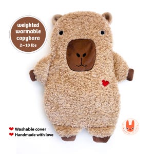 May include: A tan, fuzzy, weighted warmable capybara plush toy with a dark brown snout, small ears, and a red heart embroidered on the chest. Text reads: weighted warmable capybara 2-10 lbs, Washable cover, Handmade with love, and better critters.