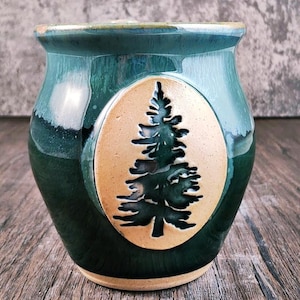 May include: A green ceramic mug with a raised design of a pine tree in a cream-colored oval. The mug has a glazed finish and a rounded shape.