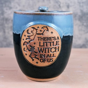 May include: A ceramic mug with a blue and black glaze. The mug features a circular emblem with the text "THERE'S A LITTLE WITCH IN ALL OF US" and a witch silhouette. The mug has a small handle on the top.