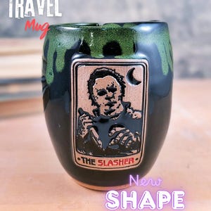 May include: Black and green ceramic travel mug with a copper-colored rectangular emblem featuring a horror movie character and the words "THE SLASHER." The mug has the words "TRAVEL Mug" and "New SHAPE" in the image.