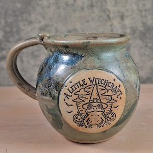 May include: A round ceramic mug with a mottled green and blue glaze featuring a circular tan medallion on the front. The medallion displays a black line drawing of a frog wearing a witch hat, flanked by two pumpkins, with the text A LITTLE WITCHCRAFT.