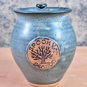 May include: A handmade, blue-green ceramic jar with a lid. The jar features a circular emblem with a tree and the words "Spooky Season." The jar has a rustic, textured appearance, perfect for Halloween or fall decor.