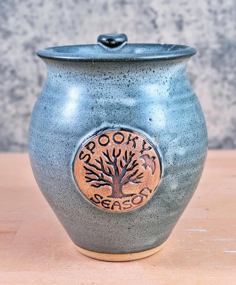 May include: A handmade, blue-green ceramic jar with a lid. The jar features a circular emblem with a tree and the words "Spooky Season." The jar has a rustic, textured appearance, perfect for Halloween or fall decor.