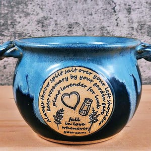 May include: A handcrafted ceramic bowl with a blue and black glaze. The bowl has two handles and a circular emblem with text and illustrations. The text reads "Always throw spilt salt over your left shoulder, keep rosemary by your garden gate, plant lavender for luck, fall in love whenever you can."