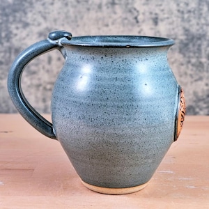 May include: A handmade ceramic mug in a speckled blue-gray glaze. The mug features a rounded body, a sturdy handle, and a decorative emblem. The base is unglazed, revealing the natural clay color. A unique piece for enjoying beverages.