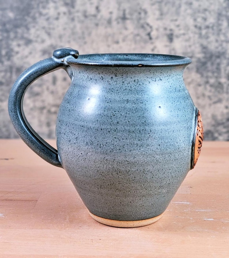 May include: A handmade ceramic mug in a speckled blue-gray glaze. The mug features a rounded body, a sturdy handle, and a decorative emblem. The base is unglazed, revealing the natural clay color. A unique piece for enjoying beverages.