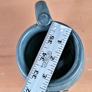 May include: A handmade, gray-green ceramic mug with a rounded handle. The mug is approximately 4 inches (10 cm) tall, as measured by a ruler. The base of the mug features a decorative, embossed design.