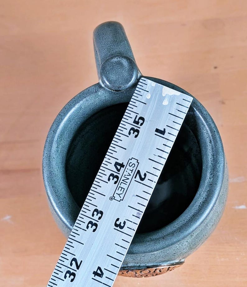 May include: A handmade, gray-green ceramic mug with a rounded handle. The mug is approximately 4 inches (10 cm) tall, as measured by a ruler. The base of the mug features a decorative, embossed design.