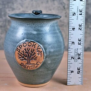 May include: A blue-grey ceramic jar with a lid, featuring a circular emblem with a tree design and the words "SPOOKY SEASON." The jar is approximately 5 inches tall.