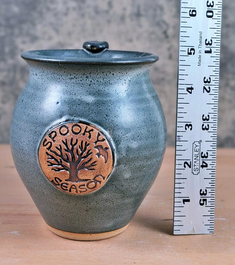 May include: A blue-grey ceramic jar with a lid, featuring a circular emblem with a tree design and the words "SPOOKY SEASON." The jar is approximately 5 inches tall.