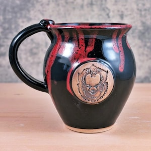 May include: Black ceramic mug with a red glaze dripping down the sides. The mug features a black handle and a circular emblem with a detailed face. The mug is a unique piece of pottery.