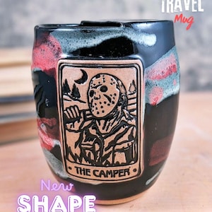 May include: Black and red ceramic travel mug with a rectangular embossed design featuring a masked figure and the text "THE CAMPER." The mug has a unique shape and the words "TRAVEL Mug" are printed on the top.