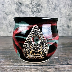 May include: Black ceramic mug with a red, white, and green glaze. The mug features a gold-colored Ouija board planchette with the words "Hello & Goodbye" on it.
