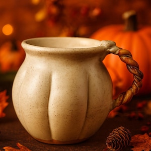 May include: A cream-colored ceramic mug with a twisted handle, set against a backdrop of pumpkins, fall leaves, and berries. The mug has a textured surface and a unique, rustic design, perfect for autumn decor.