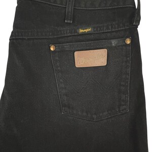 May include: Black denim jeans featuring a high-rise waistband with belt loops, a back pocket with a tan leather patch embossed with the Wrangler logo, gold-tone metal rivets, and the signature W stitching detail on the back pocket.