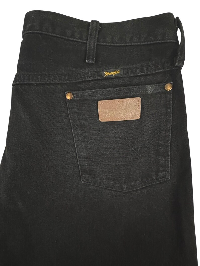 May include: Black denim jeans featuring a high-rise waistband with belt loops, a back pocket with a tan leather patch embossed with the Wrangler logo, gold-tone metal rivets, and the signature W stitching detail on the back pocket.