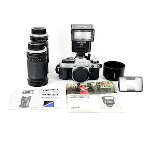 May include: A silver and black Canon AE-1 camera with a mounted flash, two black Tamron zoom lenses, a lens hood, a flash diffuser, and two owner's manuals. The camera body features a black lens cap and the text Canon AE-1 on the front.