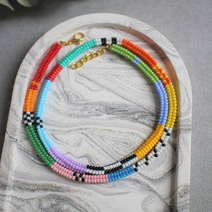 Puede incluir: Collar de cuentas multicolores con cierre dorado. El collar está hecho de pequeñas cuentas redondas en tonos de rojo, naranja, amarillo, verde, azul, morado y negro. Las cuentas están dispuestas en un patrón repetitivo.