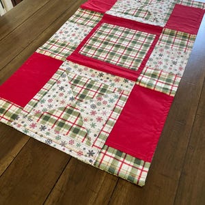 May include: A rectangular fabric table runner featuring a patchwork design of solid red panels, green and red plaid squares, and a cream-colored fabric printed with small red and green snowflakes. The quilted textile has stitched seams.