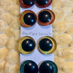 May include: A collection of four pairs of large, round, doll eyes in various colors. Each eye has a black pupil and a glittery, colored iris. The colors include red, orange, yellow, and teal. The text "Alex Marie Crochet" is written on the paper.