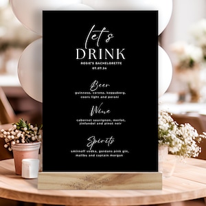 Könnte beinhalten: Schwarz-weißes Schild mit dem Text "Let's Drink" und einer Getränkekarte für einen Junggesellinnenabschied. Das Schild steht auf einem Holzständer.
