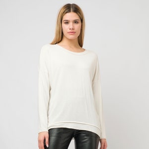 Puede incluir: Un suéter blanco de manga larga con cuello redondo y un ajuste ligeramente holgado. El suéter se lleva con pantalones de cuero negro.