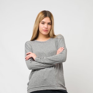 Puede incluir: Una mujer con una camisa gris de manga larga con los brazos cruzados. Está de pie frente a un fondo blanco.