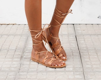 May include: A pair of tan leather gladiator sandals with laces that tie up the legs. The sandals have a flat sole and are made of a soft, supple leather.