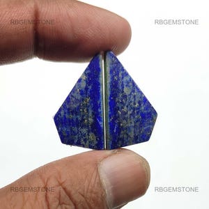May include: Two triangular, deep blue lapis lazuli gemstones with gold flecks are held between fingers. The gemstones are cut to fit together, with a thin silver line separating them. The gemstones are from RBGEMSTONE.