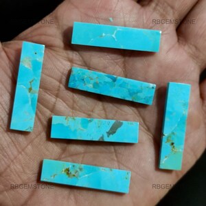 Puede incluir: Seis cabujones de turquesa, cortados en forma rectangular, de color azul verdoso con vetas marrones. Las piedras están dispuestas en una mano.