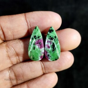 Può includere: Due pietre preziose a forma di goccia con base verde, macchiate di nero e un tocco di magenta. Le pietre sono tenute in una mano, mostrando le loro superfici lucide e i modelli naturali. Le pietre preziose sono probabilmente per la creazione di gioielli.