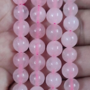 May include: Close-up of several strands of pink rose quartz beads. The beads are spherical and have a soft, translucent appearance. They are strung together with a pink cord. The text "RBGEMSTONE" is visible in the corners of the image.