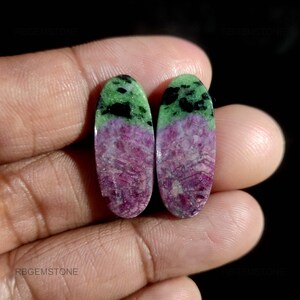Puede incluir: Dos gemas ovaladas con un degradado de color. La parte superior es verde con manchas negras, que se transforma en un tono rosa-púrpura en la parte inferior. Las gemas se sostienen en una mano sobre un fondo negro.